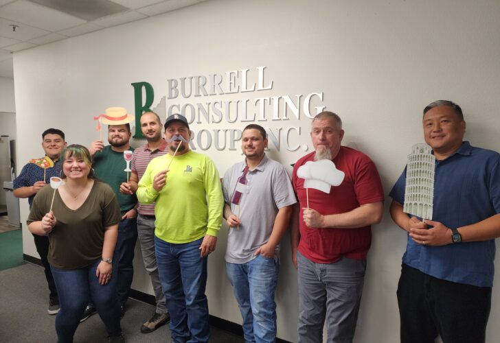 A group of eight people stands in front of a wall with the text "Burrell Consulting Group, Inc." Several individuals are holding themed props, including a chef's hat, wine glass, pizza slice, and the Leaning Tower of Pisa.
