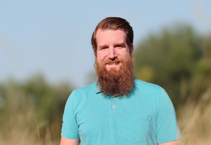 A bearded man in a teal polo shirt stands smiling in a field of tall grass under a clear blue sky, with a blurred background of trees.